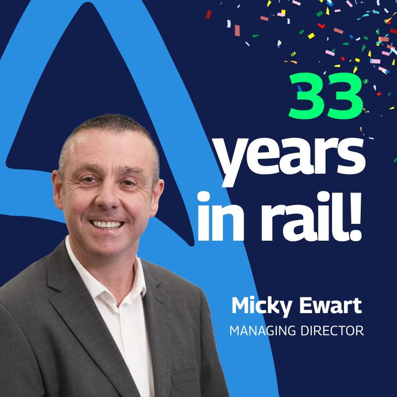 The image is a celebratory graphic highlighting Micky Ewart’s 33 years in the rail industry. It features a portrait of Micky, a Caucasian male with a friendly smile, dressed in a business suit. Behind him, there’s a large blue triangle and colorful confetti, adding a festive touch to the graphic. The text “33 years in rail!” is prominently displayed at the top, with his name and title, “Managing Director,” at the bottom. The overall color scheme is blue, white, and green, which contributes to a vibrant and professional presentation.