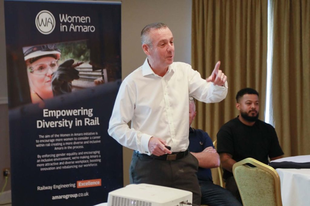 The photo captures a speaker, a Caucasian middle-aged man in a white shirt, energetically addressing an audience at a conference. Behind him, a banner for the “Women in Amaro” initiative promotes “Empowering Diversity in Rail.” The banner outlines the initiative’s goal to attract more women to the rail industry, enhancing diversity and making Amaro more inclusive. Another male attendee in the background listens intently. The setting appears to be a well-lit conference room.