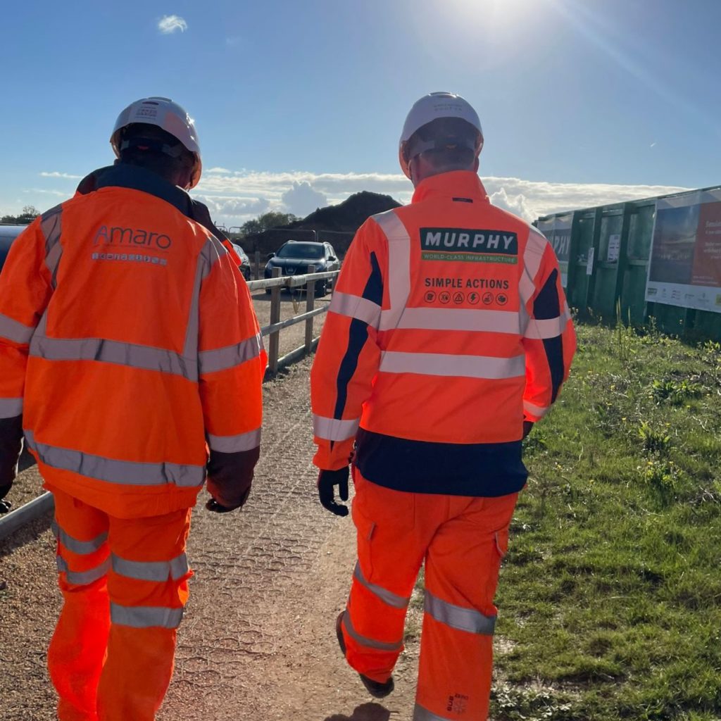 Two construction workers in high-visibility orange and navy uniforms walking on a sunlit pathway. Their jackets display the logos and names ‘Amaro’ and ‘Murphy,’ with the text ‘SIMPLE ACTIONS’ prominently featured on the back of one jacket. They are wearing white safety helmets, and the early morning sun creates a glow around them, highlighting a serene construction site environment.