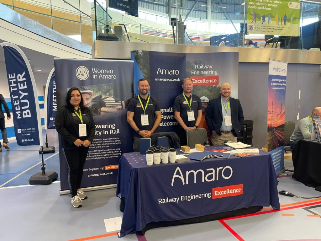 The photo depicts a group of four individuals, two men and two women, standing proudly at the Amaro exhibition booth during a conference. They are wearing business attire and lanyards, suggesting they are part of the event staff or representatives of Amaro. The booth features banners promoting the “Women in Amaro” initiative, which aims to empower diversity in the rail industry. Additional promotional materials and company branding such as “Railway Engineering Excellence” are displayed on the table and around the booth, which is located in a spacious, modern exhibition hall.