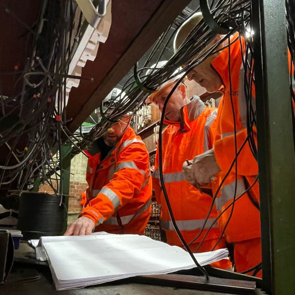 Three railway workers in high-visibility orange safety gear examining documents and inspecting a complex array of overhead cables in an industrial setting.