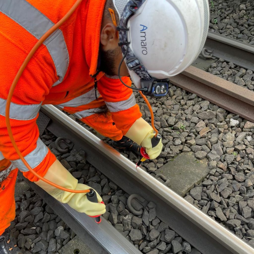 Close-up of a railway worker wearing a high-visibility orange suit and safety helmet branded 'Amaro,' attentively inspecting or repairing railway tracks, focusing on securing components with specialised gloves.