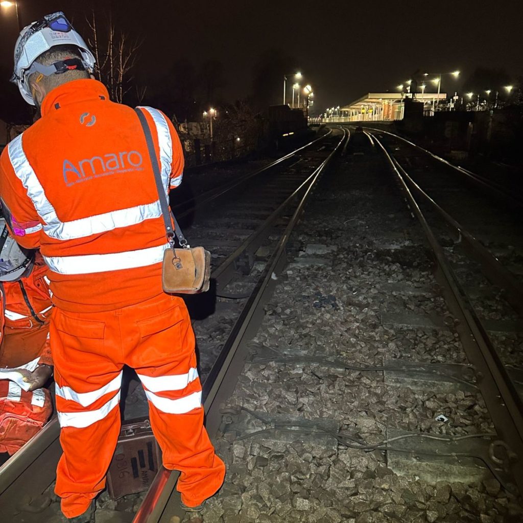 A railway worker in a high-visibility orange uniform, branded with the Amaro logo, is seen from behind, standing on the tracks at night. The scene is illuminated by ambient lighting and shows railway tracks stretching into the distance, leading towards a well-lit station. The worker, equipped with a helmet and a bag, appears to be engaged in track inspection or maintenance duties.