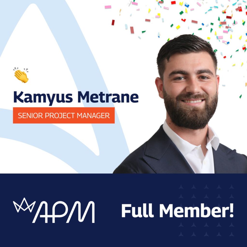 The image celebrates Kamyus Metrane's achievement of becoming a Full Member of the Association for Project Management (APM). It features a portrait of Kamyus, a bearded man with a friendly smile, dressed in a formal blue suit. Above his image, the text "Kamyus Metrane, Senior Project Manager" is prominently displayed. The APM logo is at the bottom with the words "Full Member!" The background includes a light blue peak graphic with confetti, symbolising celebration.